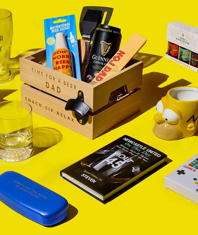 A Father’s Day gift display with a wooden crate filled with beer and snacks, surrounded by mugs, a glass, novelty socks, a book, cards and small gifts on a bright yellow background.